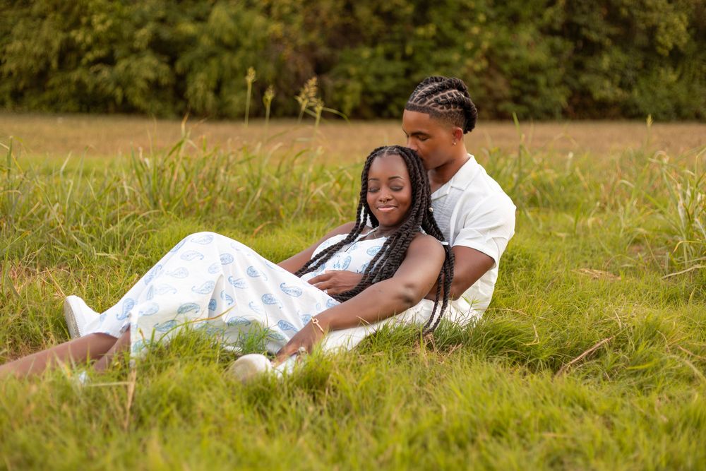 Couple sitting together in a lush green field, woman leaning back against man in matching white outfits.