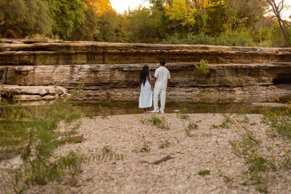 Couple standing together facing a rocky cliff by a sandy riverbank surrounded by lush green trees.