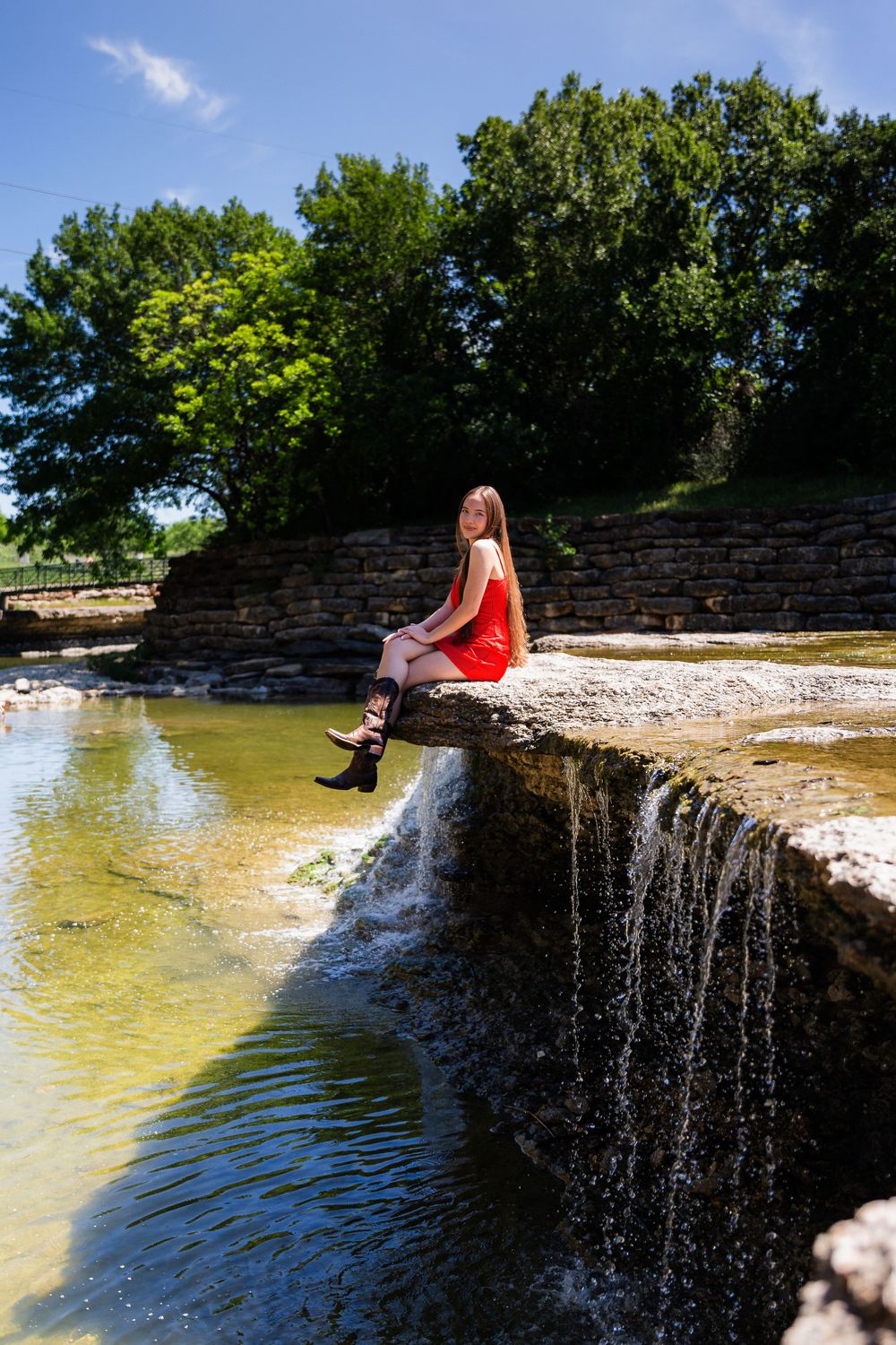 A young woman in a red dress and black boots sits on a stone ledge above a small waterfall in a sunny park.