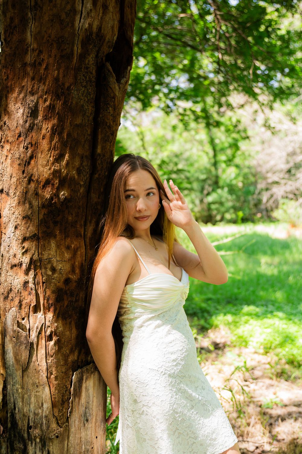 Young woman in white sundress leaning against large tree trunk in lush green park setting.