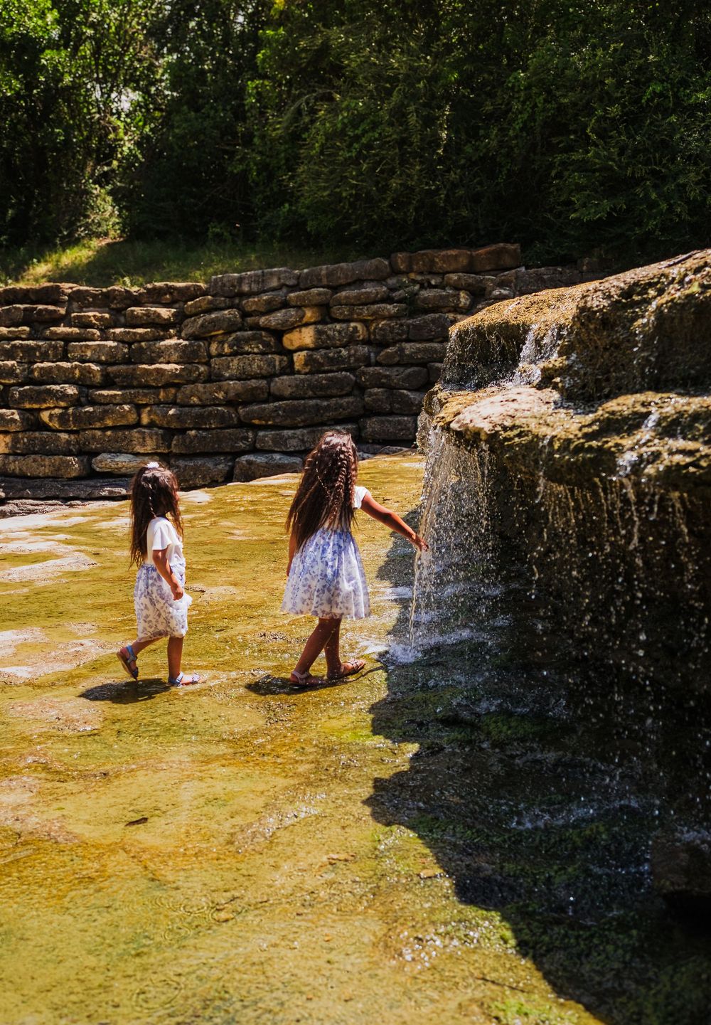 Two young girls in white dresses wade near a small waterfall beside a stone wall surrounded by lush green trees.