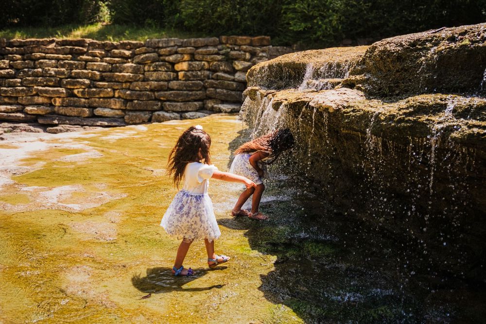 Two children playing near a small waterfall and shallow pool surrounded by stone walls on a sunny day.