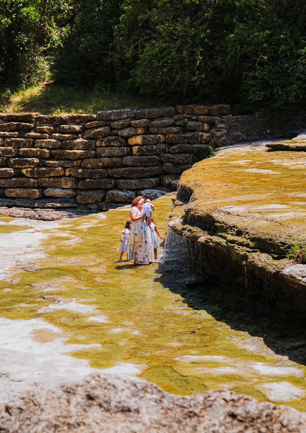 A mother and child wade in a shallow creek surrounded by limestone rocks and lush green trees.