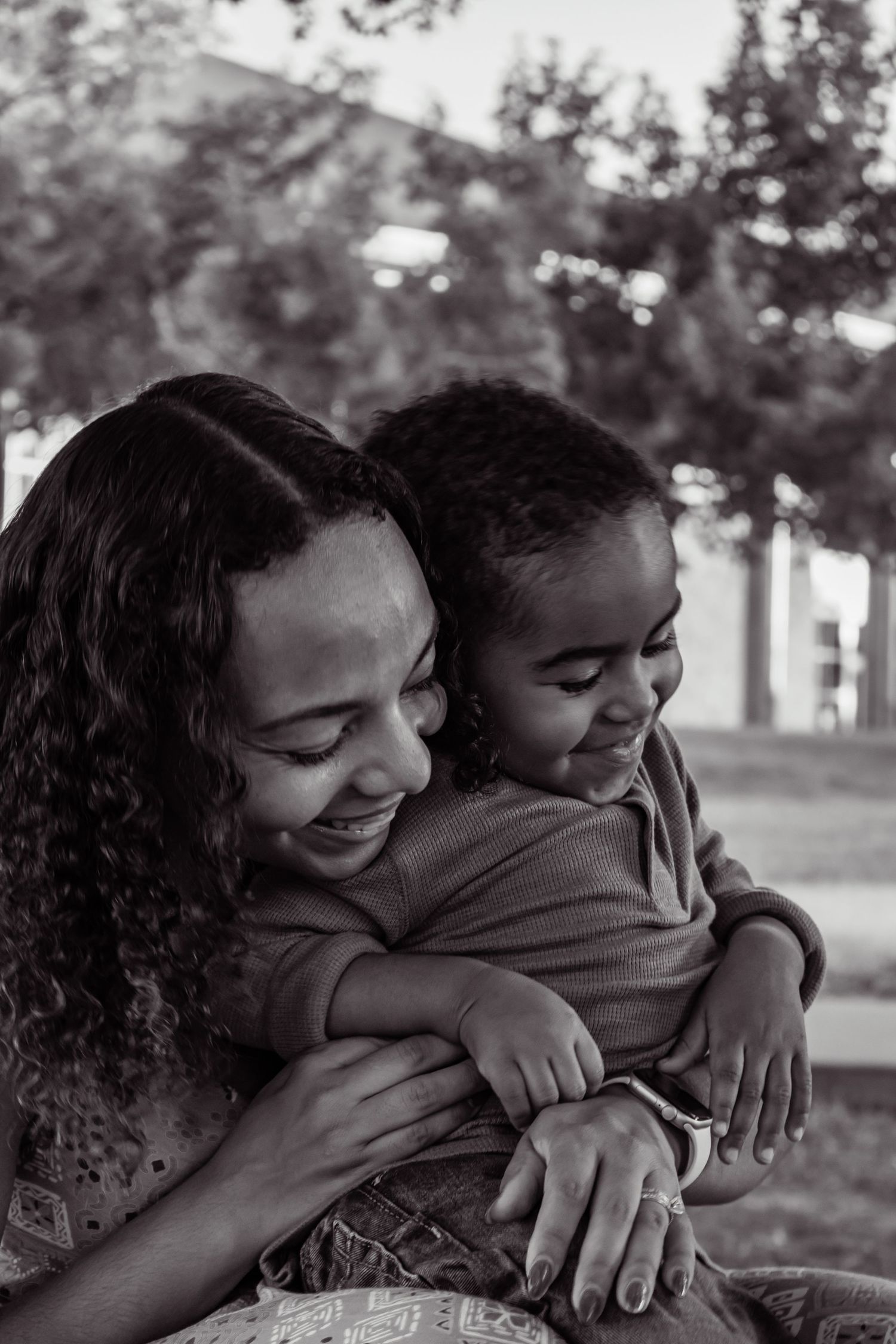 Black and white photo of a smiling mother hugging her young child outdoors in a park setting.