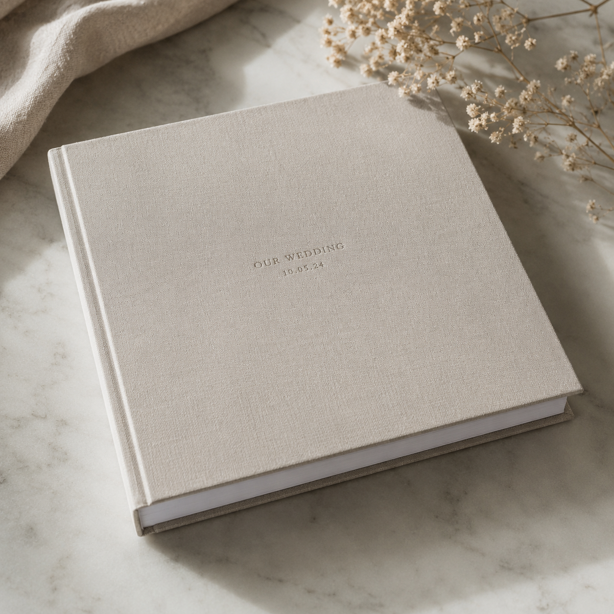 Elegant linen-covered wedding photo album with embossed text, displayed on marble surface with dried flowers.