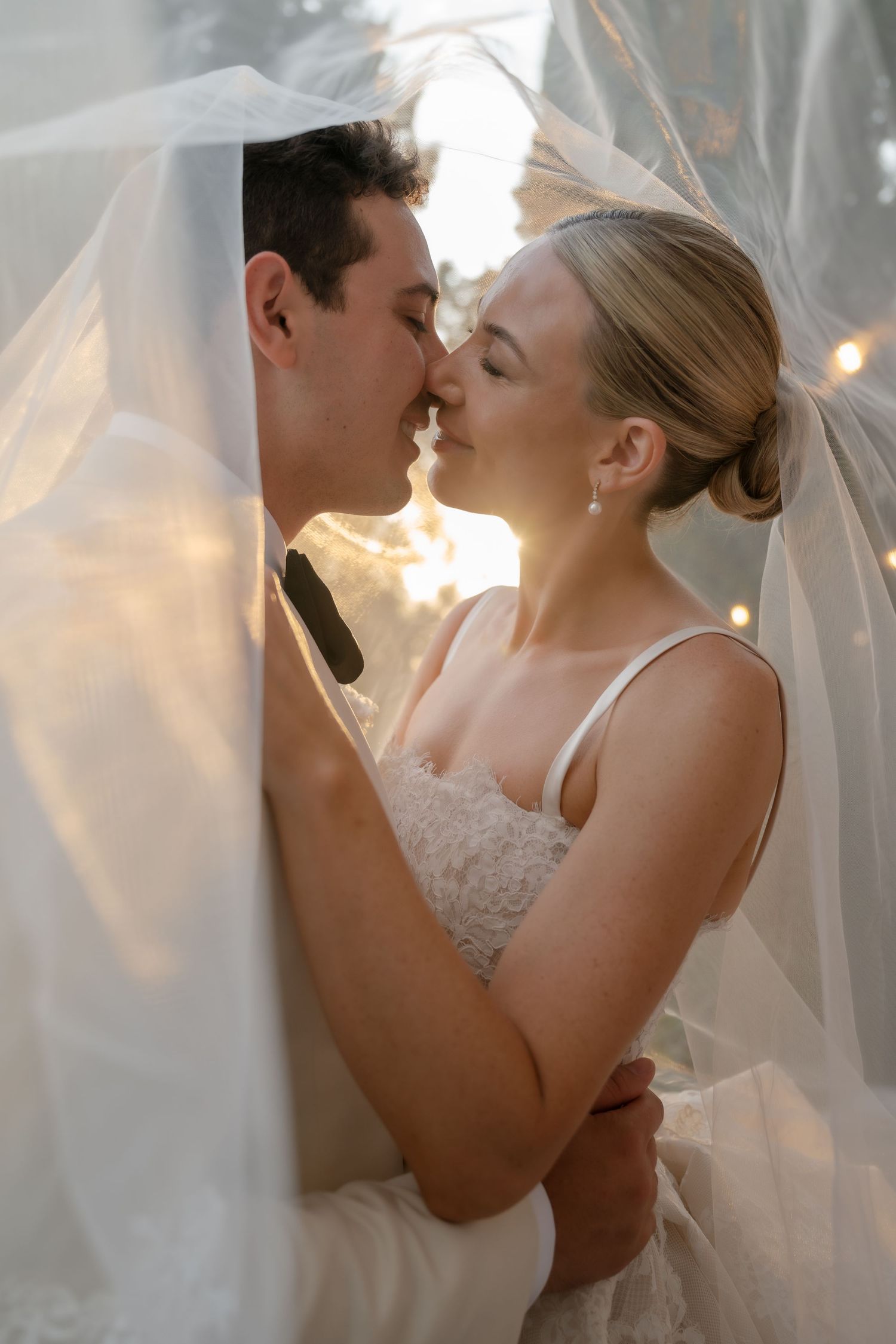 A bride and groom share a romantic kiss under a sheer wedding veil with soft golden backlight.