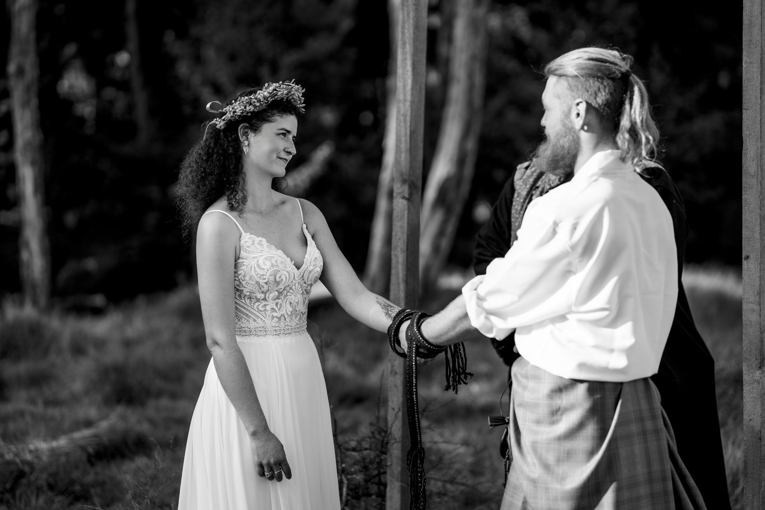 Black and white photo of a bride and groom holding hands during an outdoor woodland wedding ceremony.