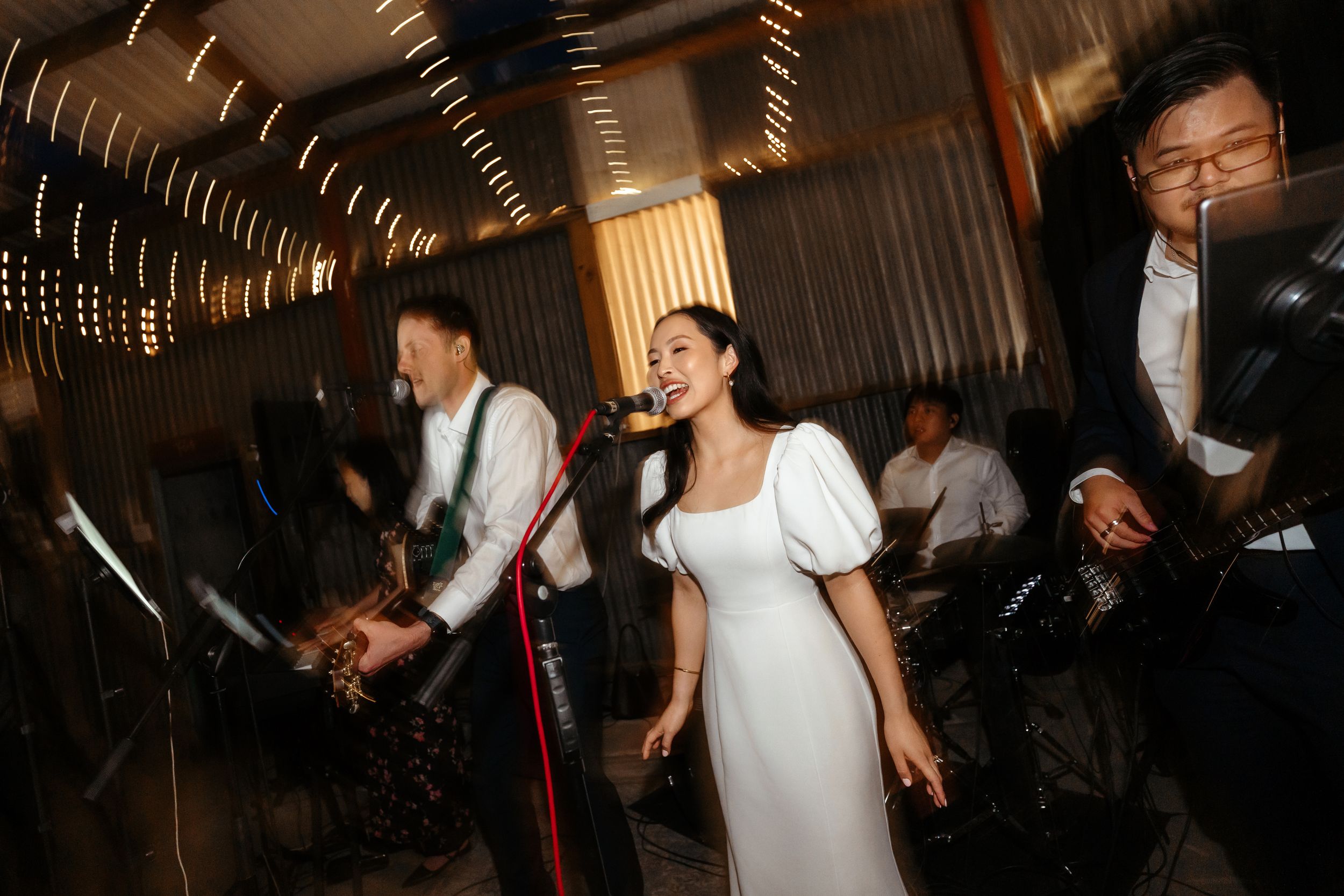 Woman in white dress singing into microphone with band members performing at lively indoor event or wedding reception.