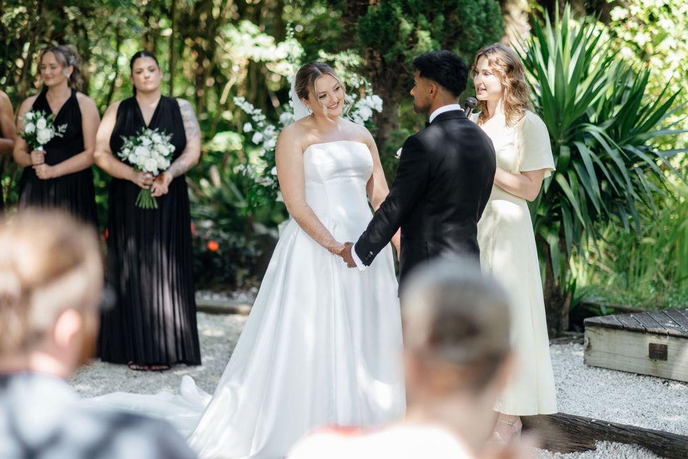 Bride and groom hold hands during outdoor wedding ceremony, with bridesmaids in black dresses and lush greenery backdrop.