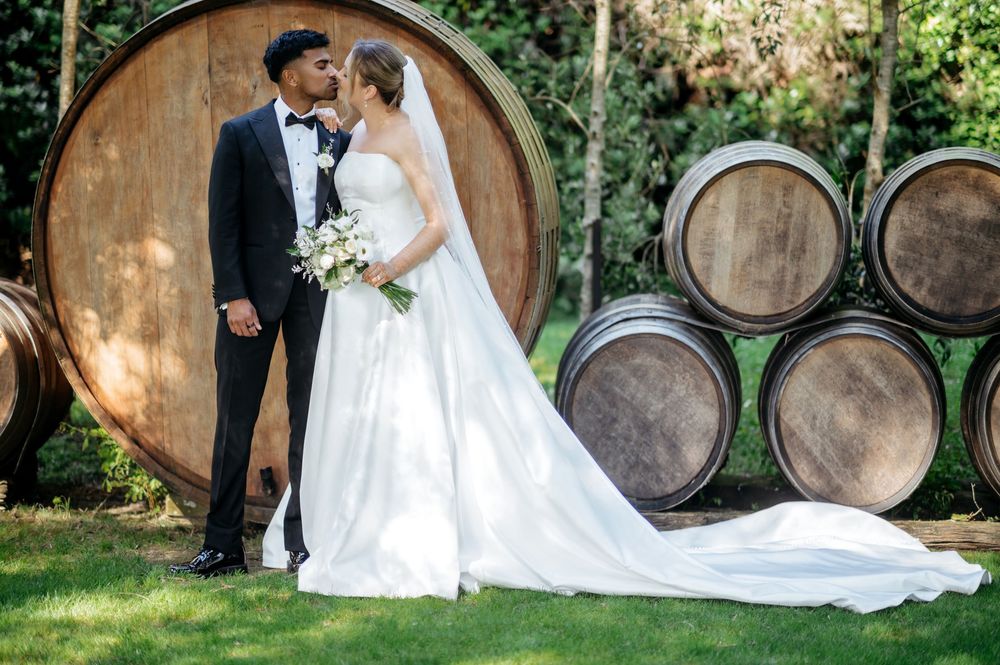 Bride and groom share a romantic kiss in front of large wooden wine barrels at a winery wedding venue.