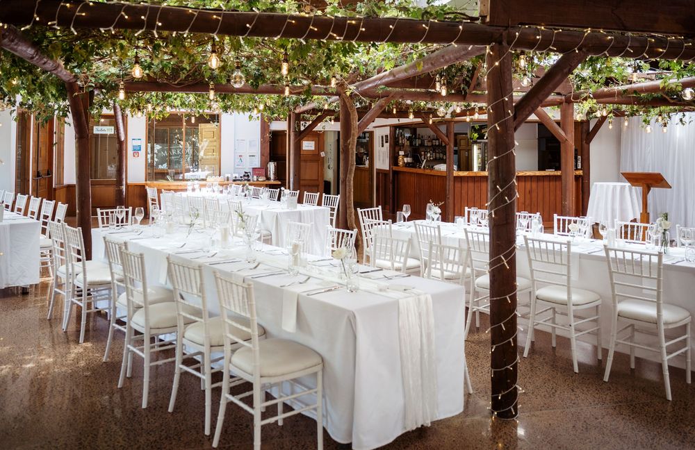 Elegant outdoor wedding reception with white chairs, long tables, white linens, and greenery draped pergola with fairy lights.