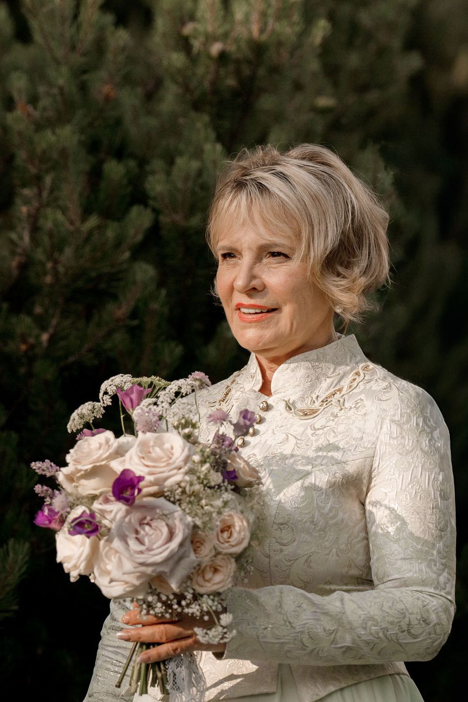 Elegant mature bride in white lace dress holding a bouquet of pink and purple roses outdoors.