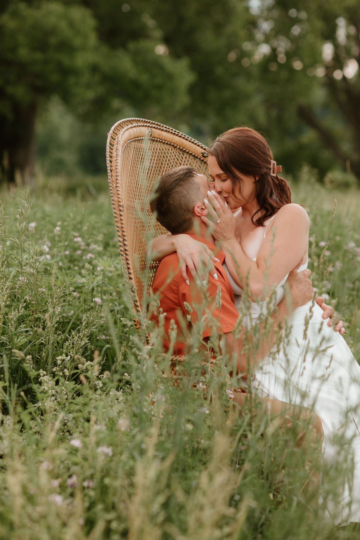 A couple shares a romantic almost-kiss in a wildflower meadow, seated in a wicker chair, dressed in orange and white.