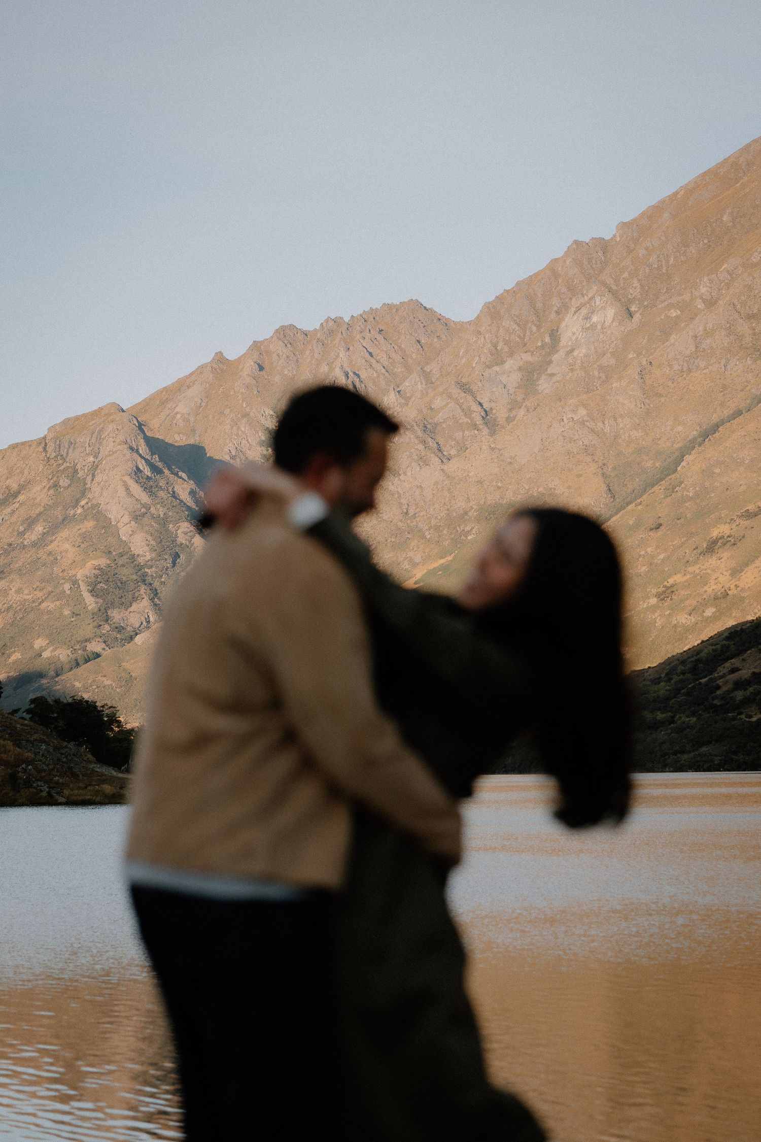 Couple embracing near a serene mountain lake at golden hour, with dramatic rocky peaks reflected in calm water.