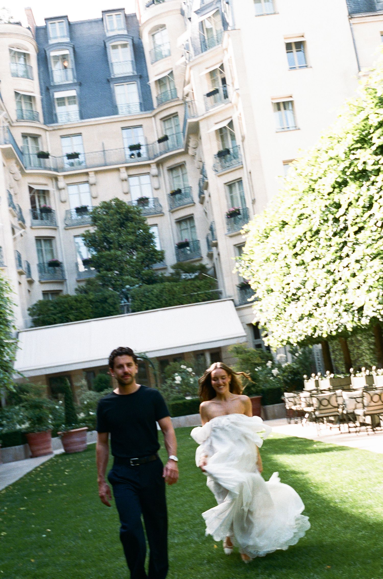 A couple walks joyfully through a lush garden courtyard of a classic Parisian hotel, woman in flowing white dress.
