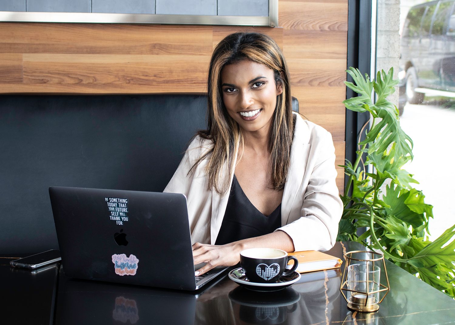 Professional woman smiling at laptop in modern cafe with coffee cup and green plant nearby.