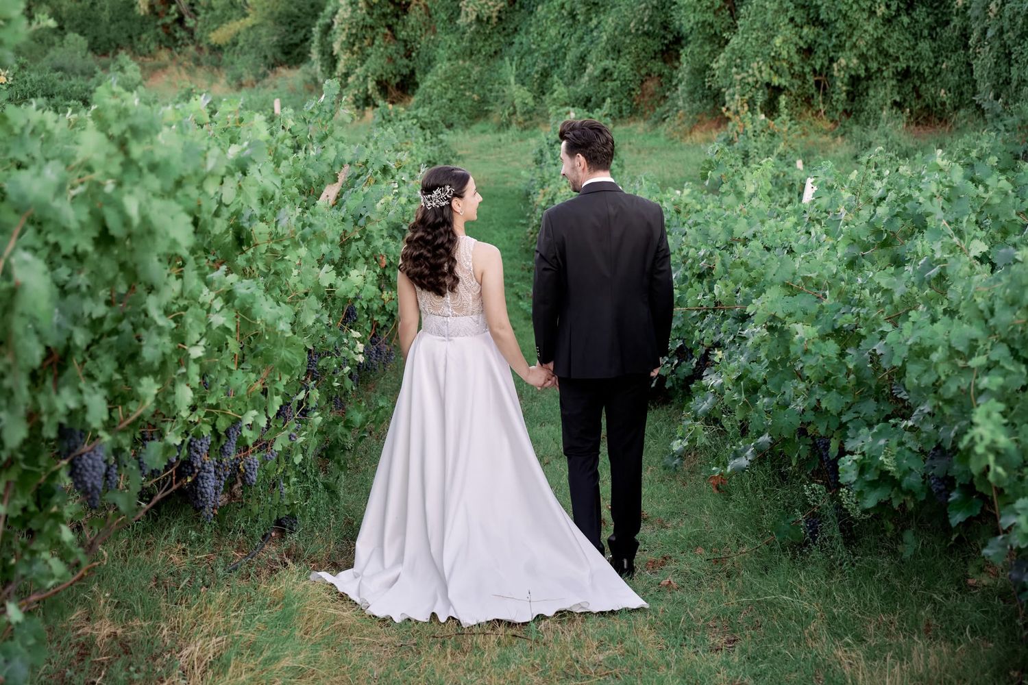 Back view of a couple in wedding attire walking hand-in-hand through a lush green field.