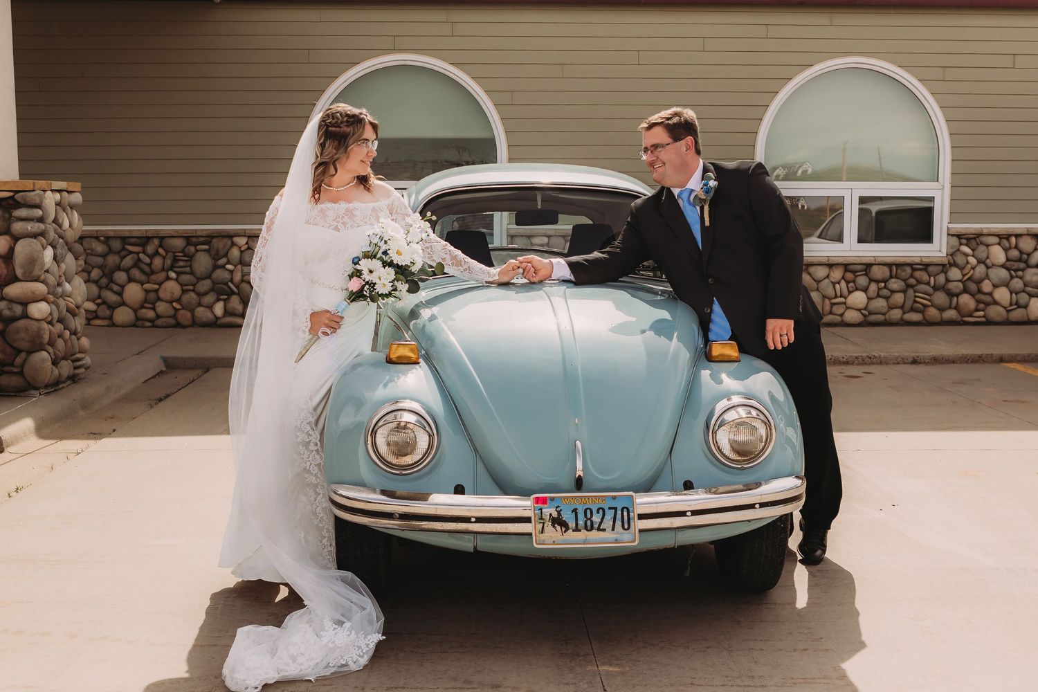 Bride and groom hold hands over a vintage light blue VW Beetle on their wedding day outdoors.