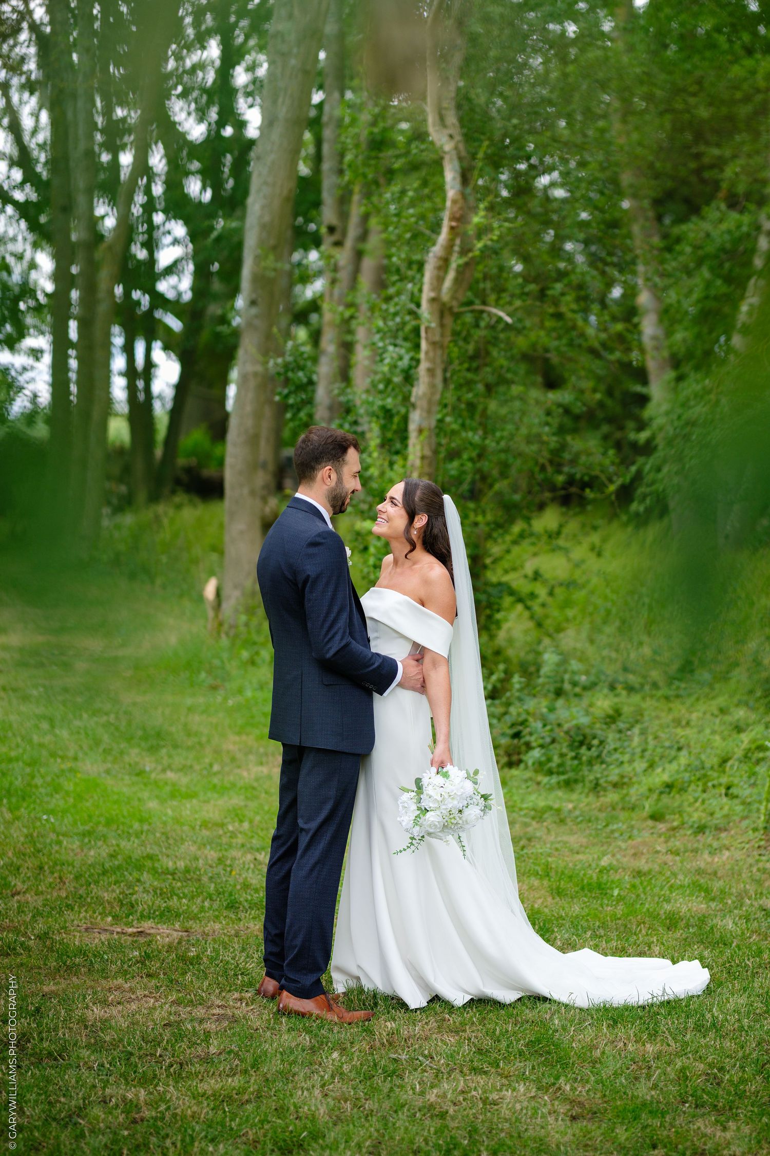 Bride and groom share a romantic moment in lush green woodland setting, bride wearing white off-shoulder gown with veil.