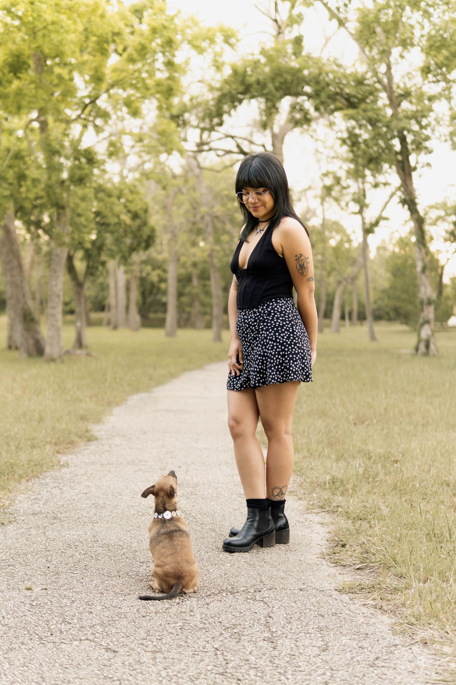 Young woman in black top and floral skirt standing on a park path with a small brown dog.