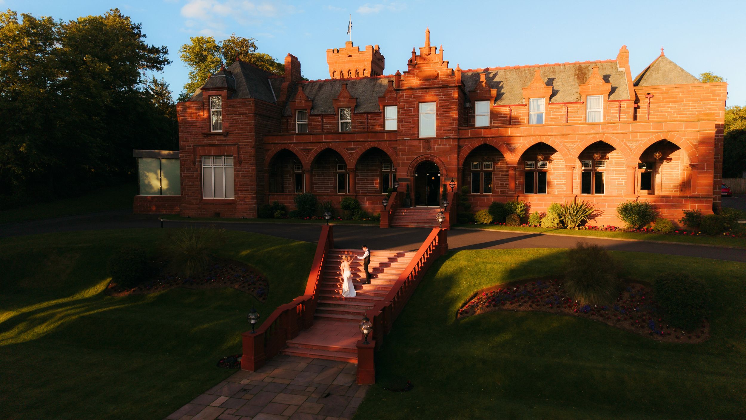 Bride in white dress descends ornate red brick castle stairs at golden hour sunset.