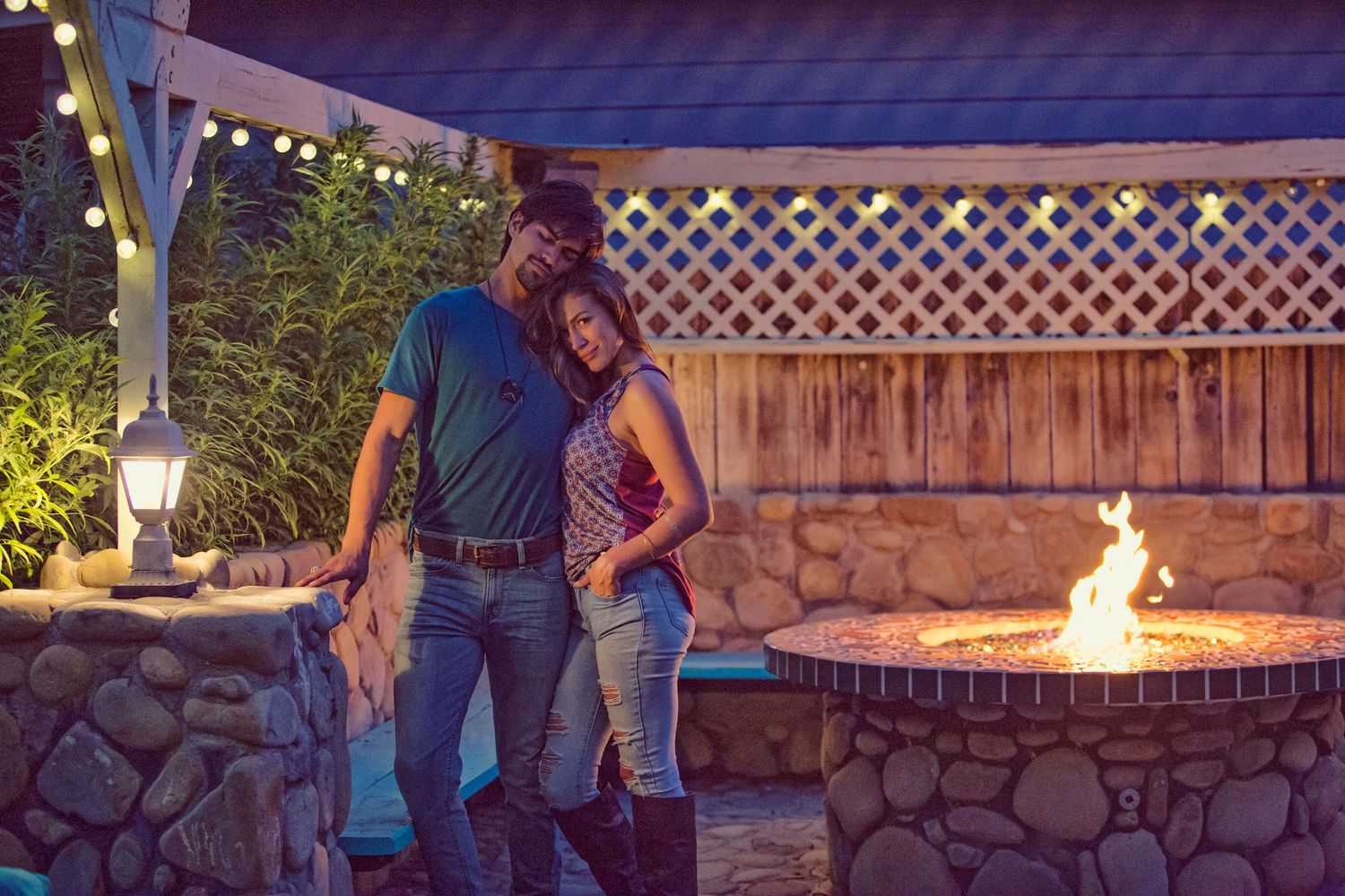 Couple embracing outdoors near a stone fire pit at night with string lights and lattice fence backdrop.