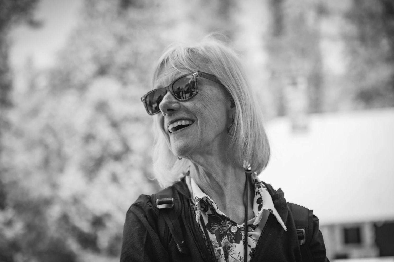Elderly woman with blonde hair and sunglasses smiling outdoors in black and white photo.
