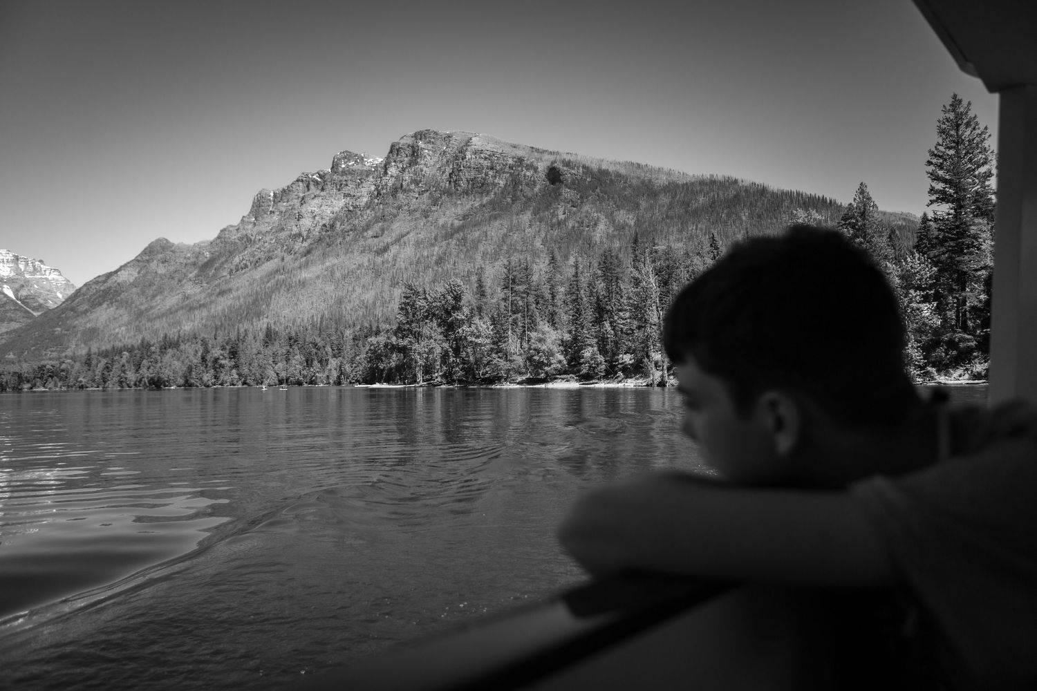 Black and white photo of a person gazing at a serene mountain lake surrounded by pine trees.