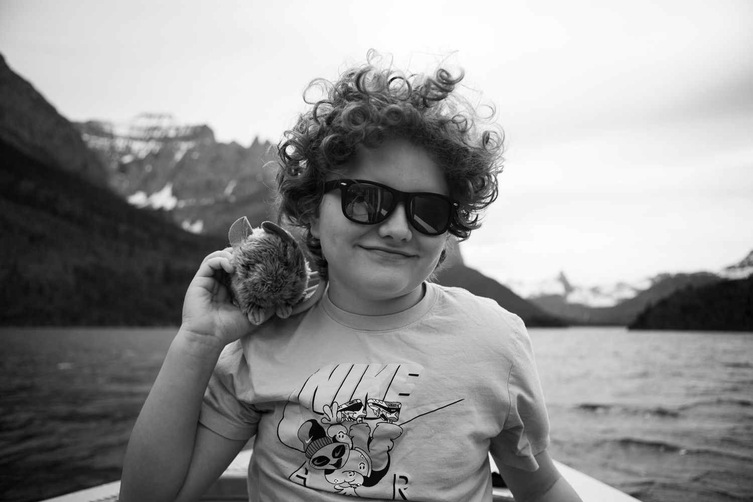 Curly-haired child wearing sunglasses and Nike shirt holds a shell near a mountain lake on a boat.