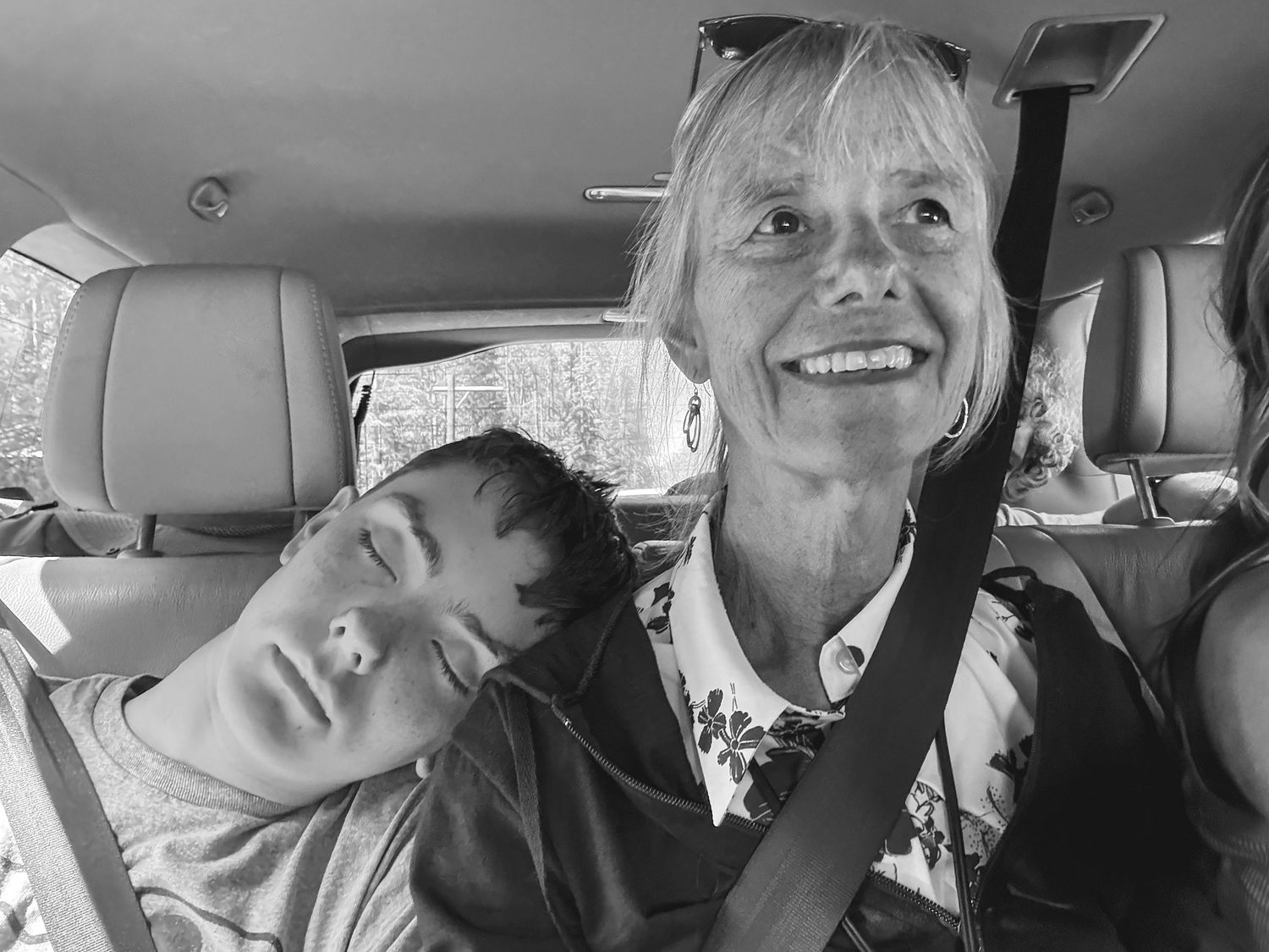 Black and white photo of a smiling woman and sleeping child in a car, both wearing seatbelts.