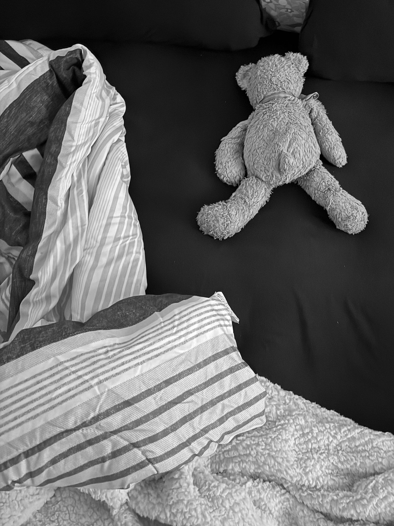 Black and white photo of a teddy bear on a dark bed with rumpled striped bedding.