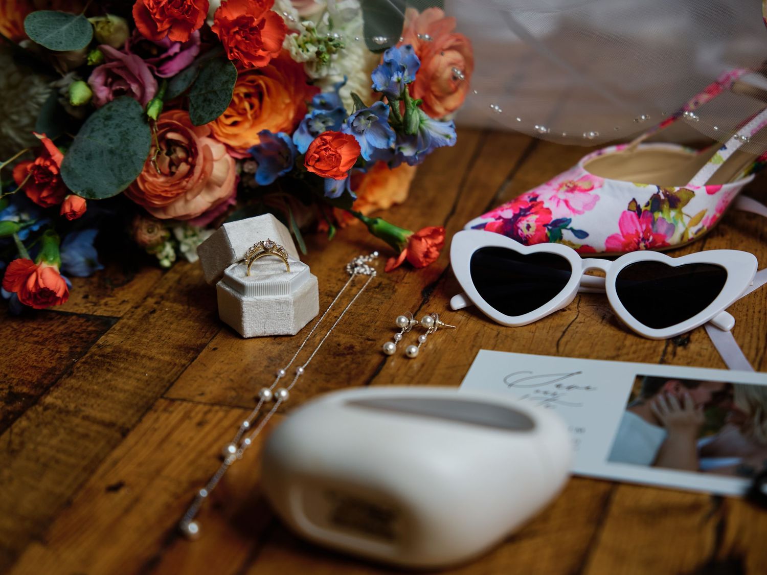 Colorful wedding flat lay with floral bouquet, heart sunglasses, ring, pearls, AirPods case, and heels on wood.