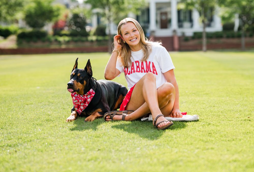 Young woman in Alabama shirt sits smiling on green grass beside a Doberman dog wearing a red floral bandana.