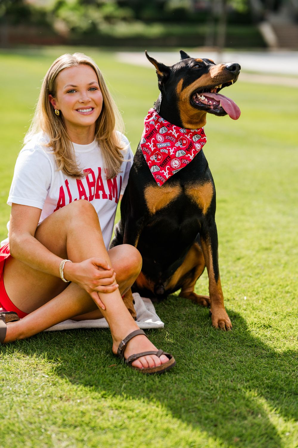 Smiling blonde woman in Alabama shirt sits on grass beside a Doberman wearing a red Alabama bandana.