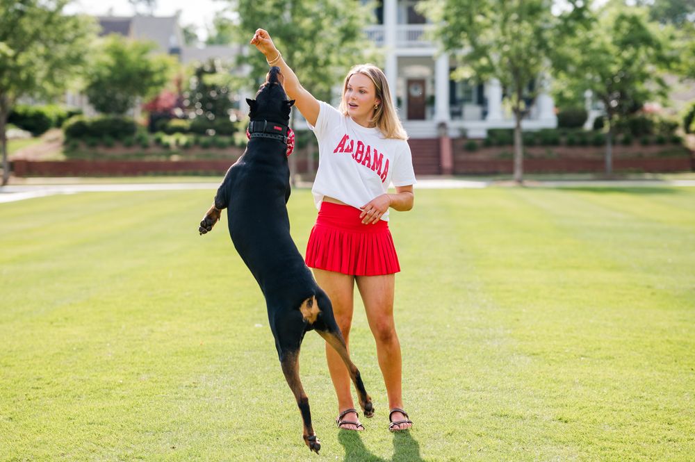 Woman in Alabama shirt and red skirt plays fetch with black dog on green lawn near white building.