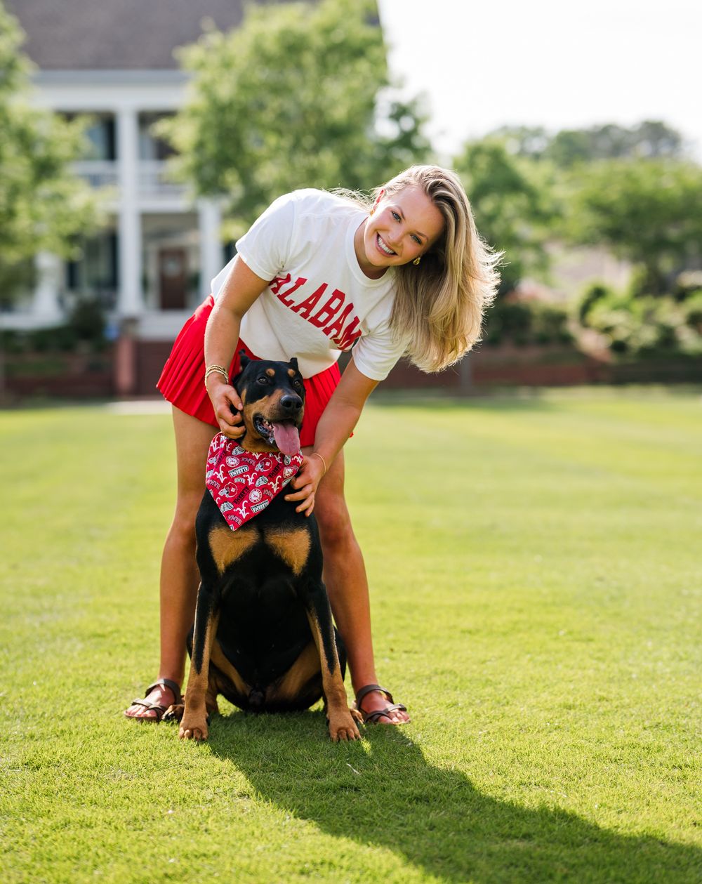 Young woman in Alabama shirt poses with a Doberman wearing a red bandana on a green lawn.