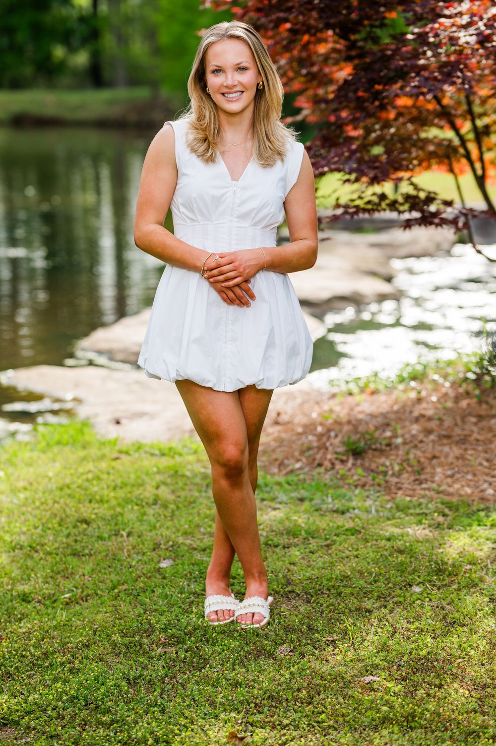 Smiling blonde woman in a white sleeveless mini dress standing by a serene pond in a lush garden.