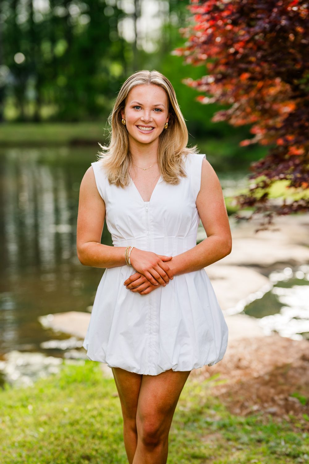 Young blonde woman in white mini dress smiling outdoors near a pond with lush green and red foliage background.