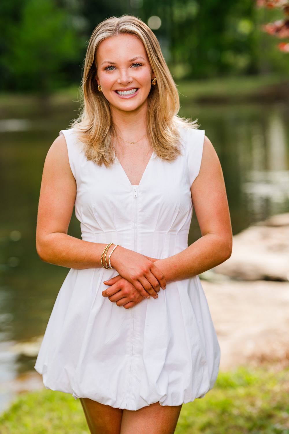 Young blonde woman smiling in white sleeveless dress, posing outdoors near a pond with green trees in background.