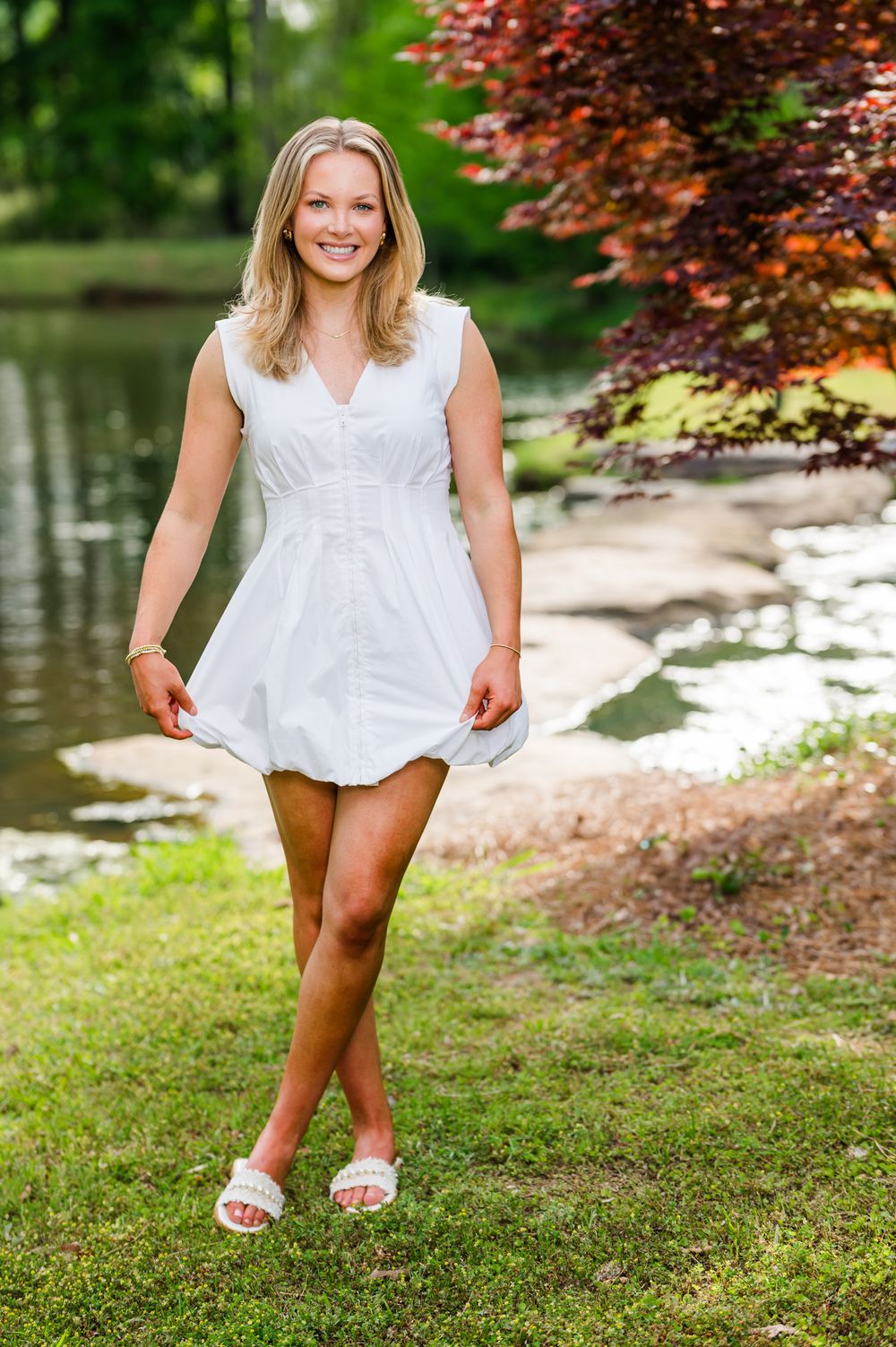 Smiling blonde woman in white dress standing by a pond in a lush green park with colorful trees.