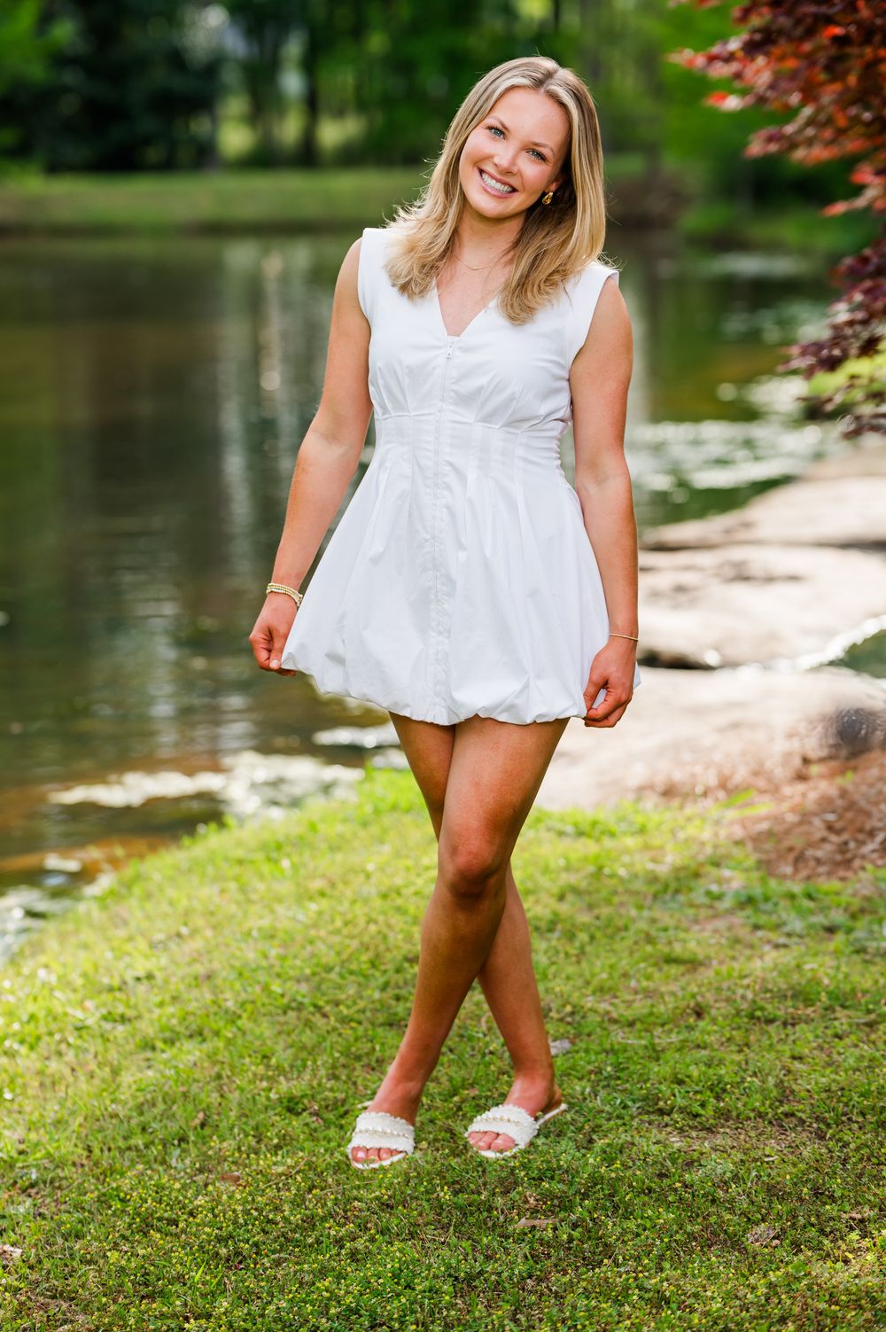 Young blonde woman smiling in white mini dress and sandals, standing by a serene pond in a lush green park.