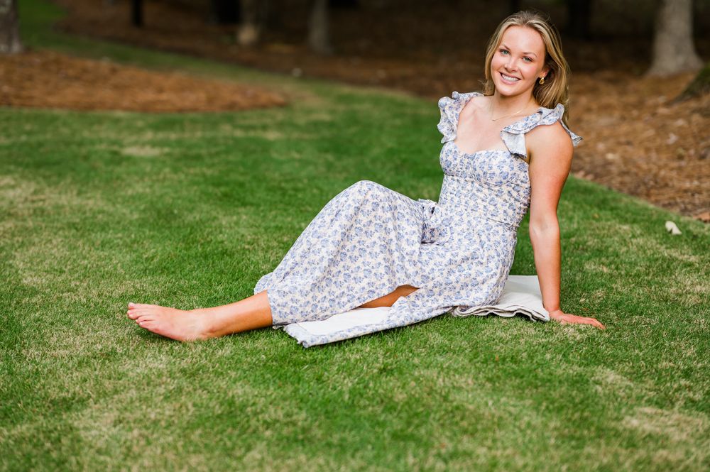 Young woman in floral sundress sitting barefoot on lush green grass, smiling outdoors in a park setting.