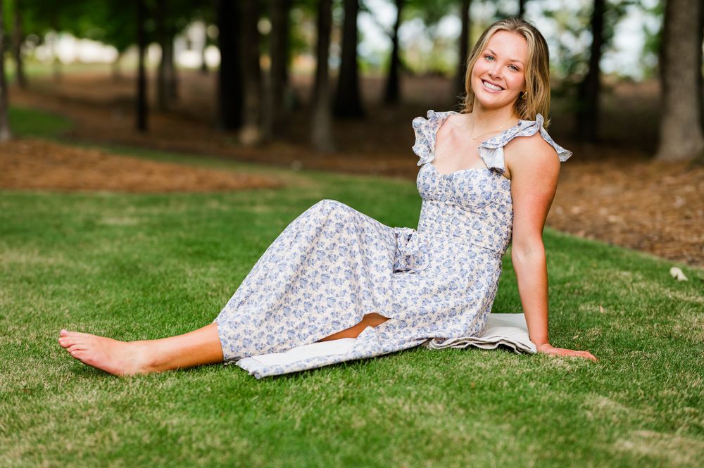 Smiling young woman in floral sundress sitting barefoot on green grass in a sunny park setting.