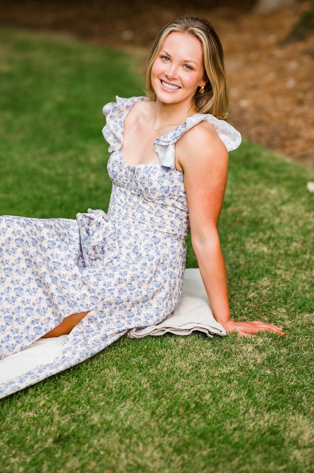 Smiling woman in floral sundress sitting on green grass outdoors in a relaxed pose.