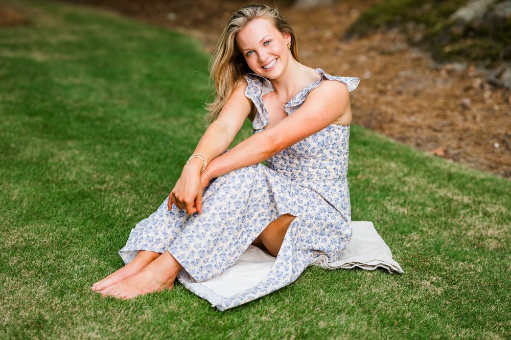 Smiling blonde woman in floral midi dress sitting barefoot on green grass outdoors.
