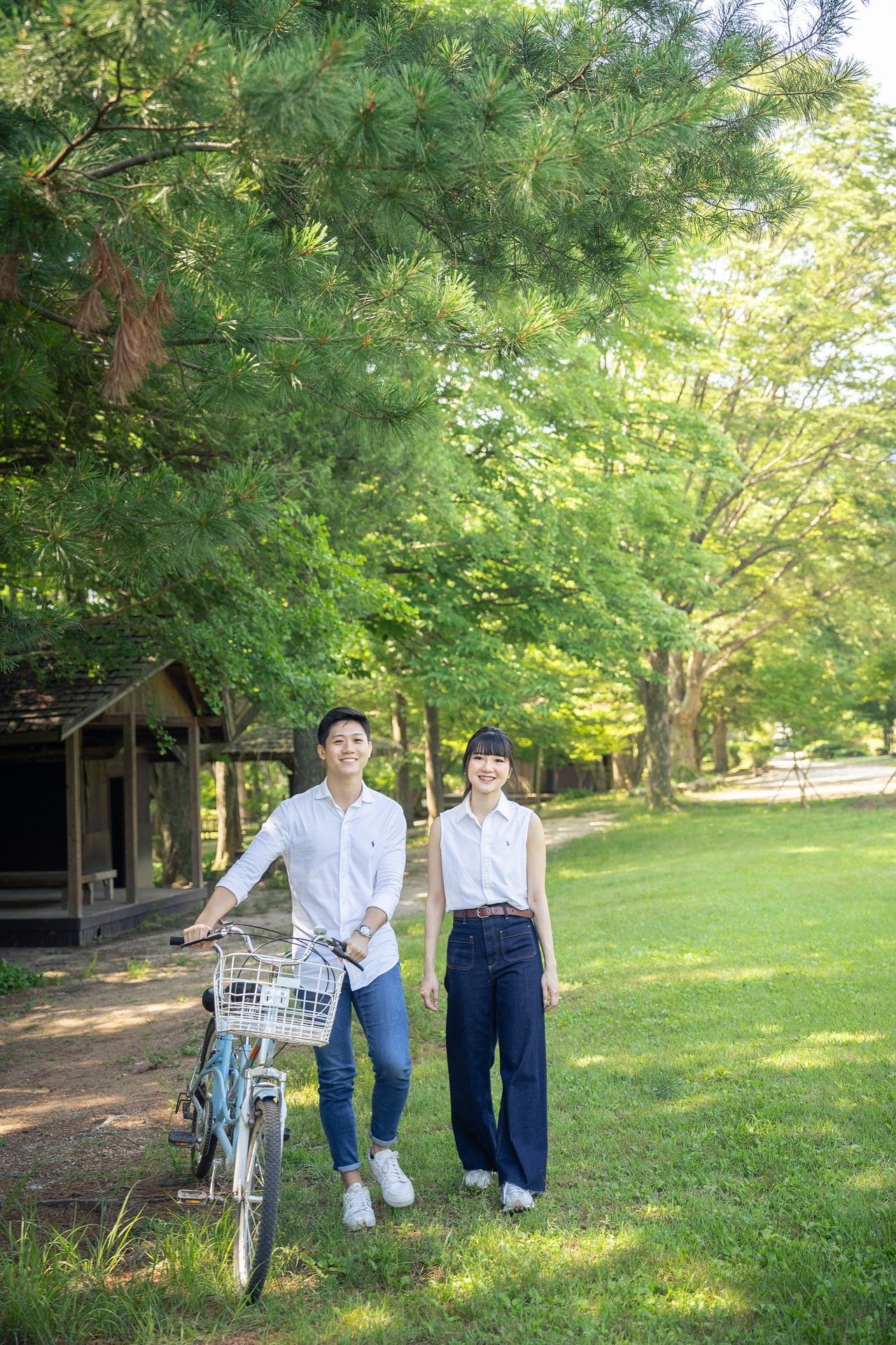 Young couple smiling and walking with a bicycle in a lush green park on a sunny day.
