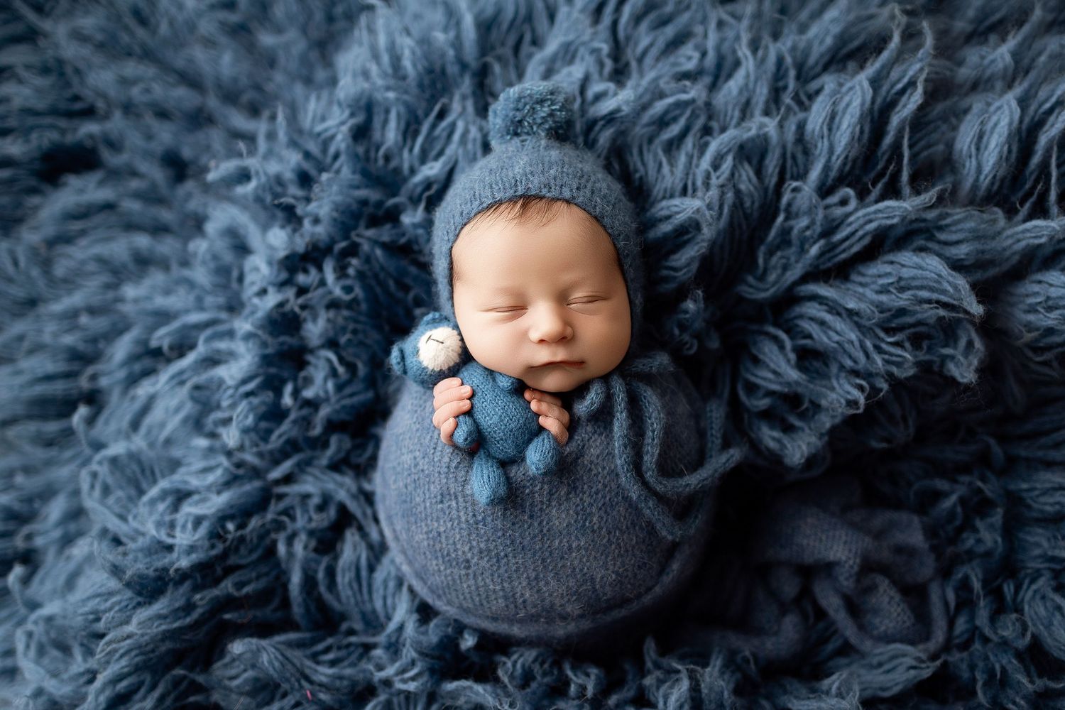 Newborn baby sleeping peacefully wrapped in blue knit blanket and hat on fluffy blue fur backdrop.