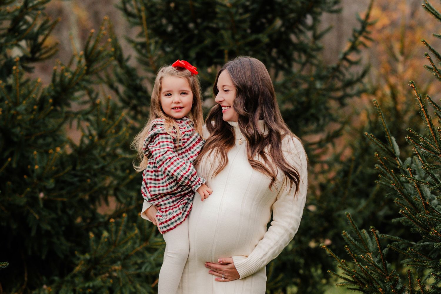Pregnant woman in white dress holding toddler girl in plaid dress among evergreen trees at a Christmas tree farm.