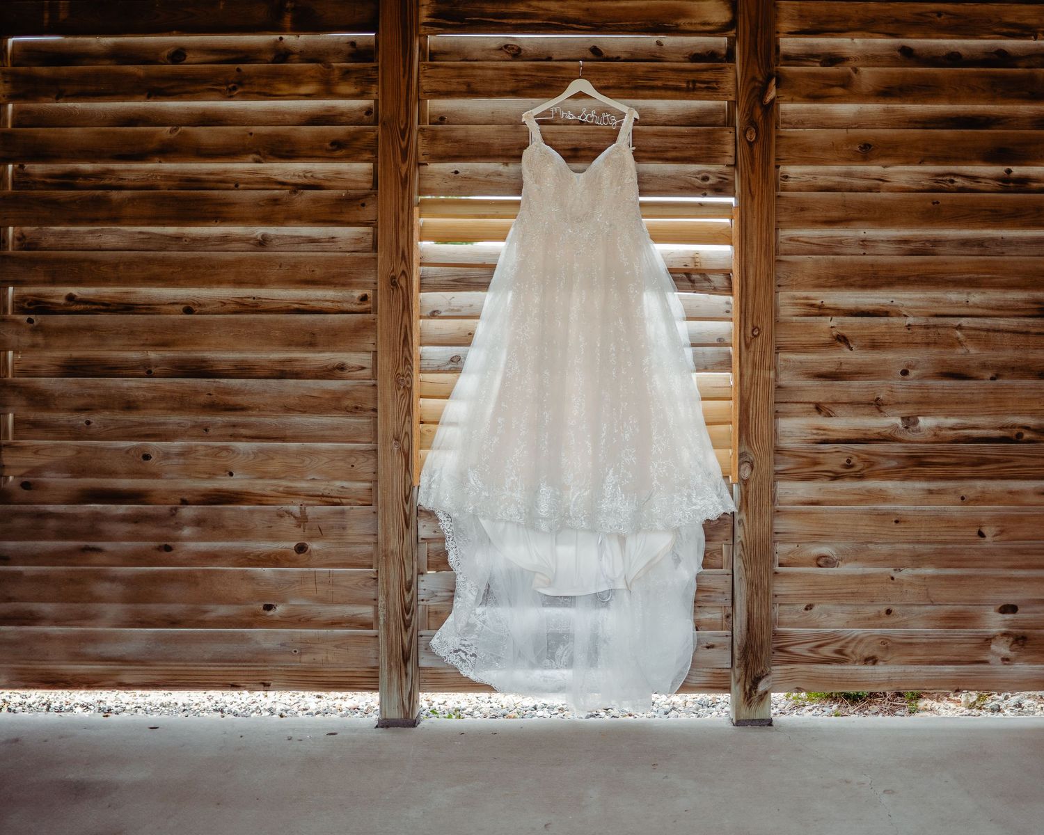 Elegant white lace wedding dress hanging on a rustic wooden barn wall, showcasing intricate details and flowing layers.