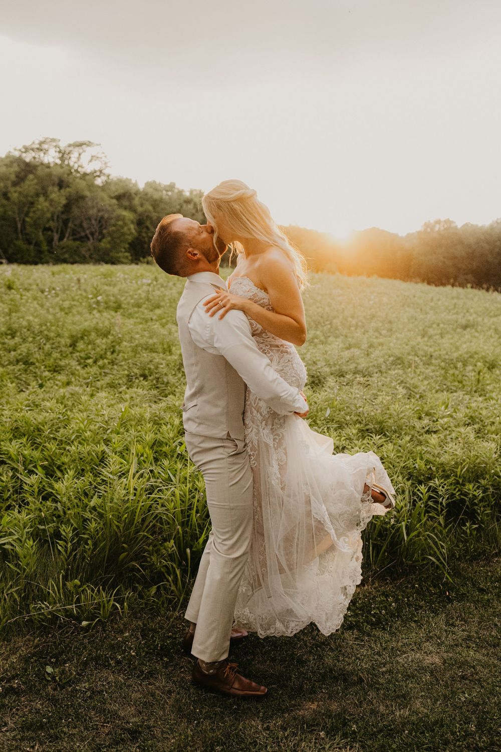 Groom lifts bride in lace wedding dress at golden hour in a lush green field.